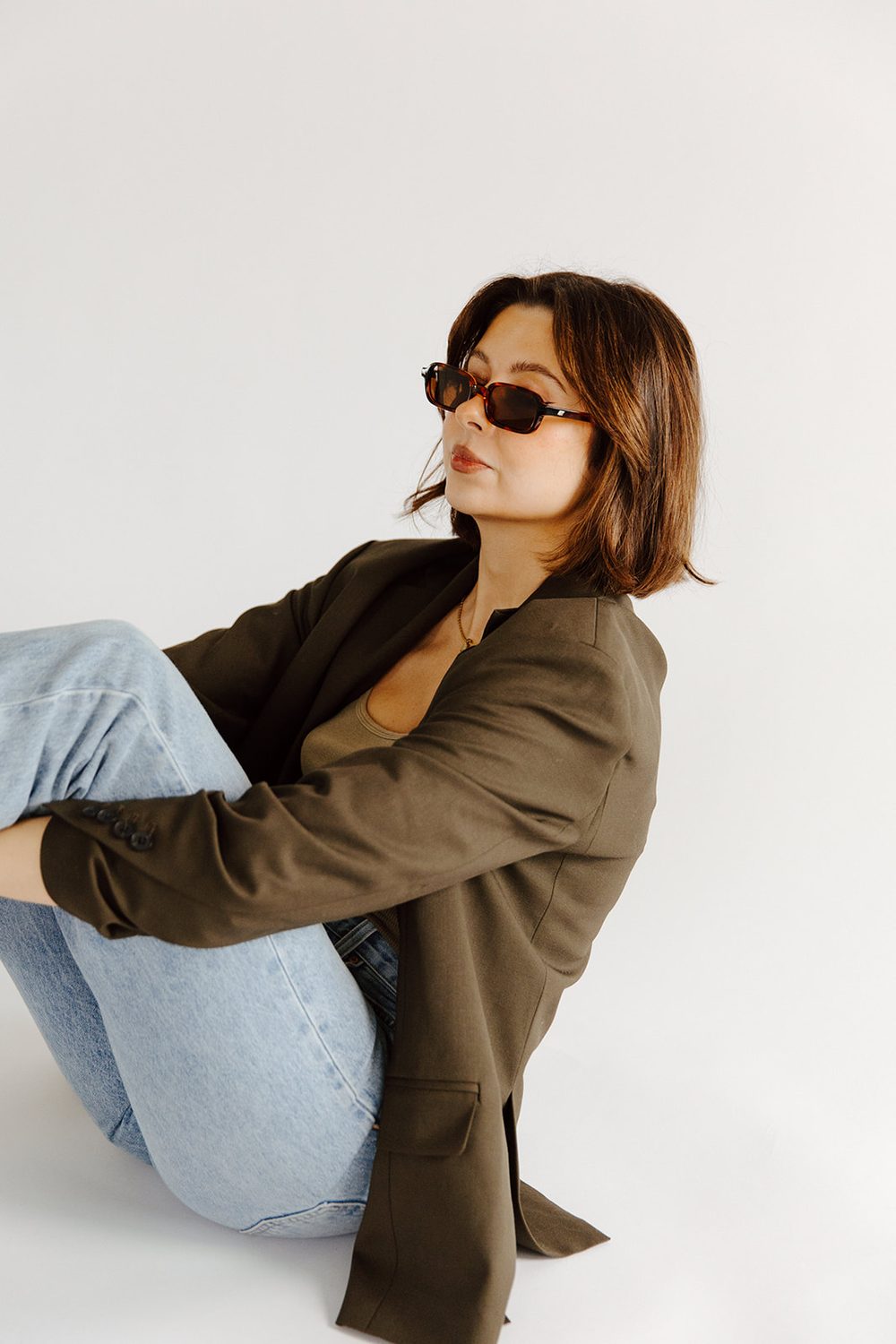 Woman in an olive blazer and light denim sitting on a white studio floor, wearing tortoiseshell sunglasses and looking off to the side