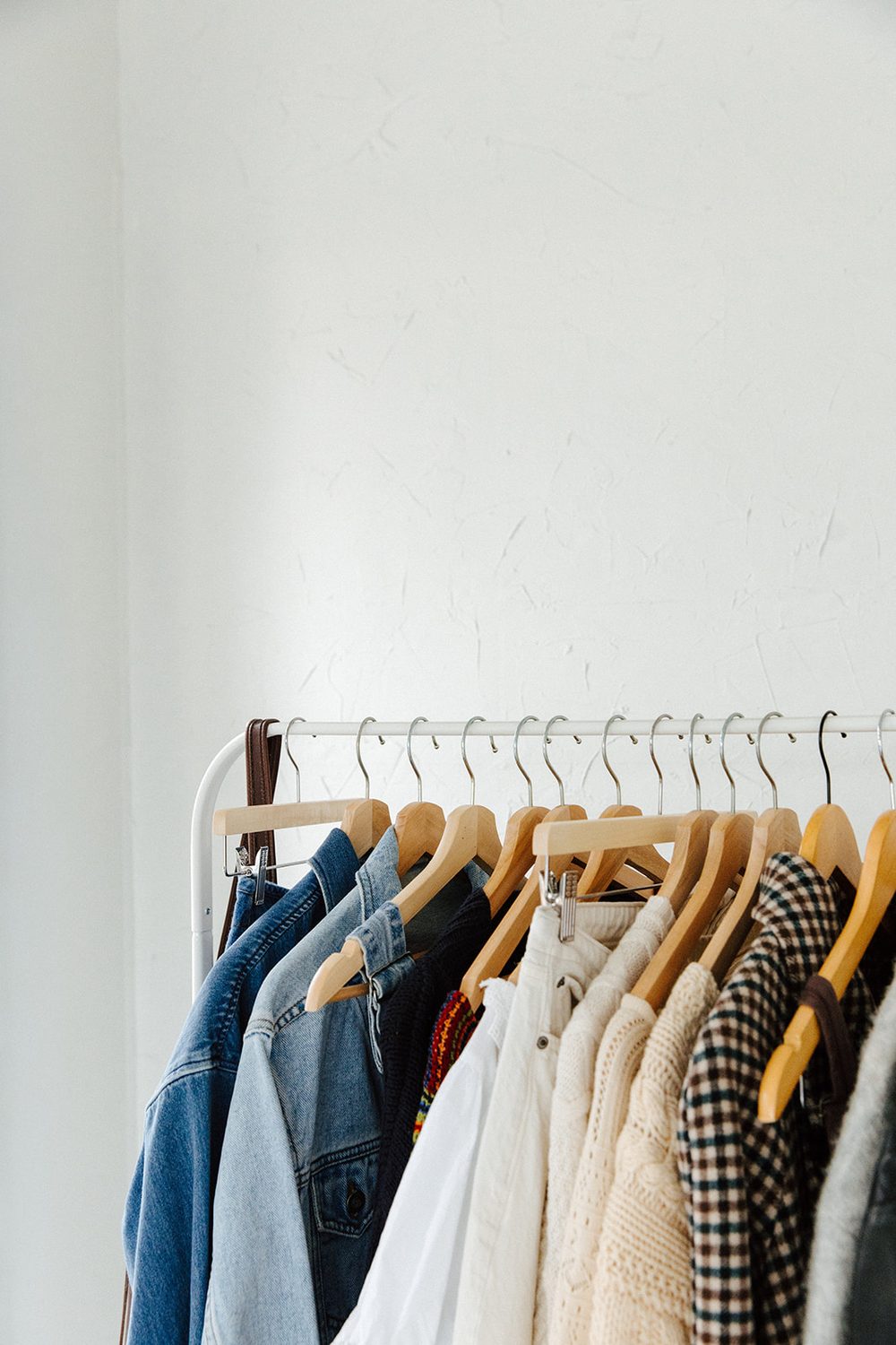 A white clothing rack with denim jackets, cream knits and a houndstooth coat on wooden hangers