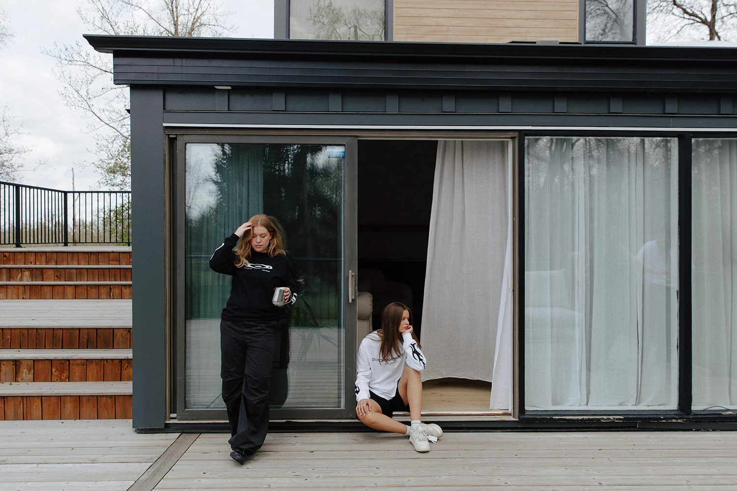 Two women outside a dark modern house, one standing with a mug and one seated in the open sliding door