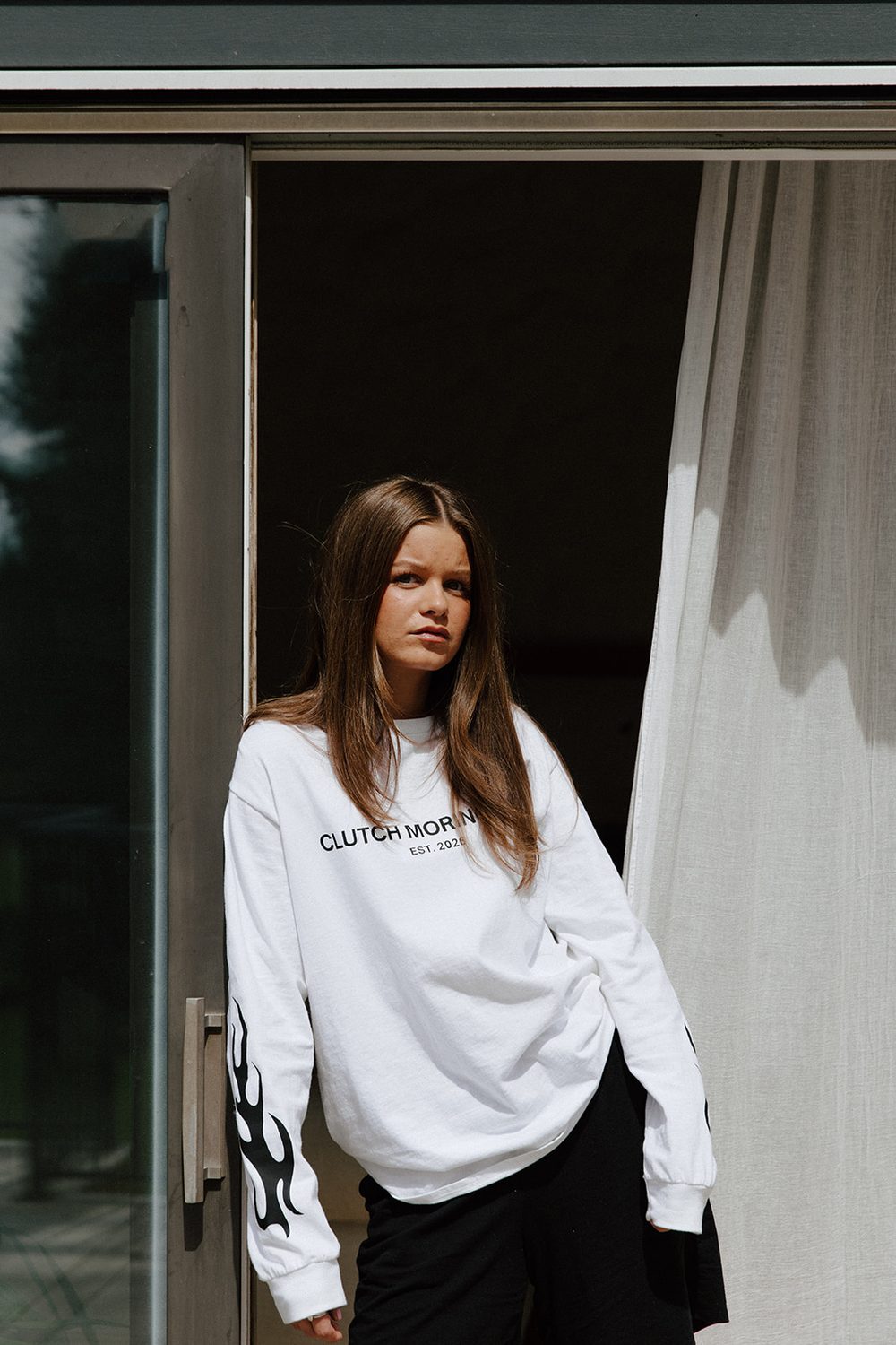 Woman in a white tee and black shorts standing in an open glass doorway beside a pale curtain