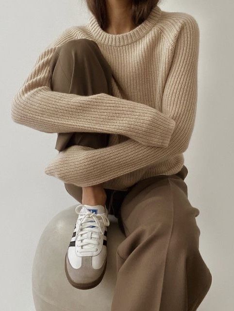Woman in a beige knit sweater and brown trousers hugging her knee, in white sneakers