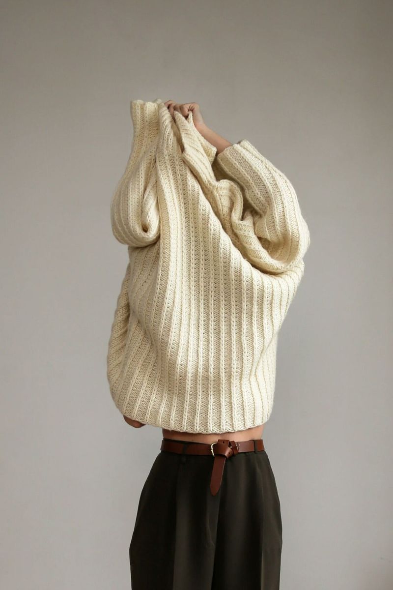 Person pulling a cream ribbed sweater over their head against a grey wall
