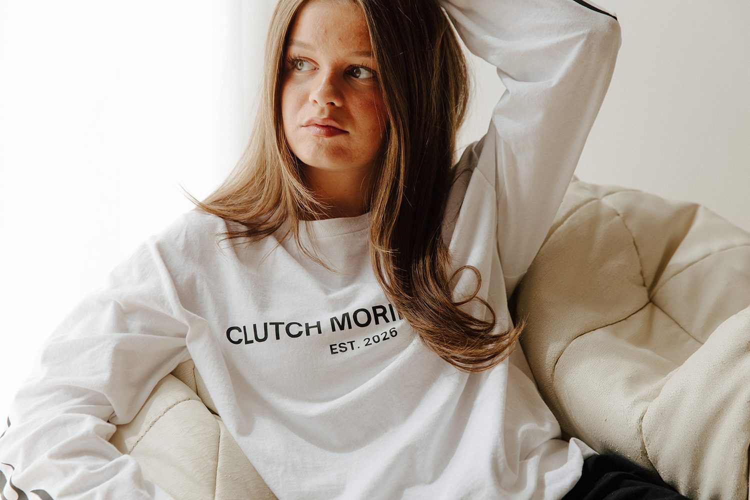 Woman in a white long-sleeve tee leaning back in a cream chair by a bright window, looking off to the side