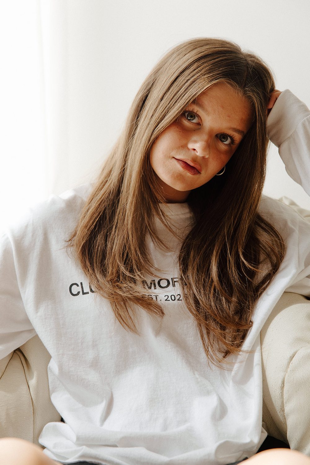 Woman with long brown hair in a white long-sleeve tee, hand in her hair, looking at the camera