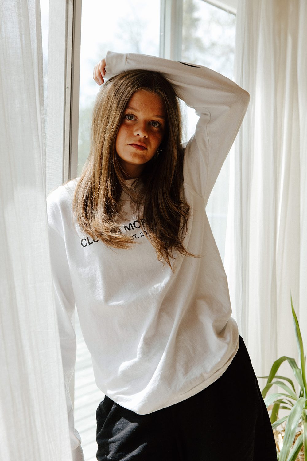 Woman in a white long-sleeve tee standing between sheer curtains at a window, arm raised over her head